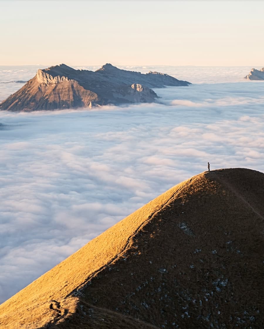 A lone hiker stands on a sunlit ridge above a sea of golden-hour clouds — gear earned, summit within reach.